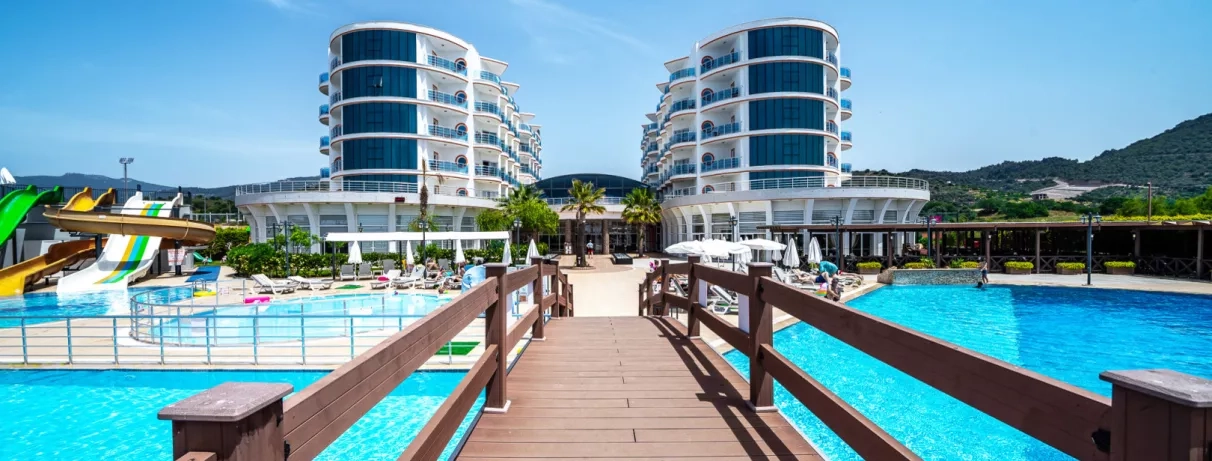 Un ponton en bois mène vers deux bâtiments d'hôtel modernes entourés de grandes piscines bleues.