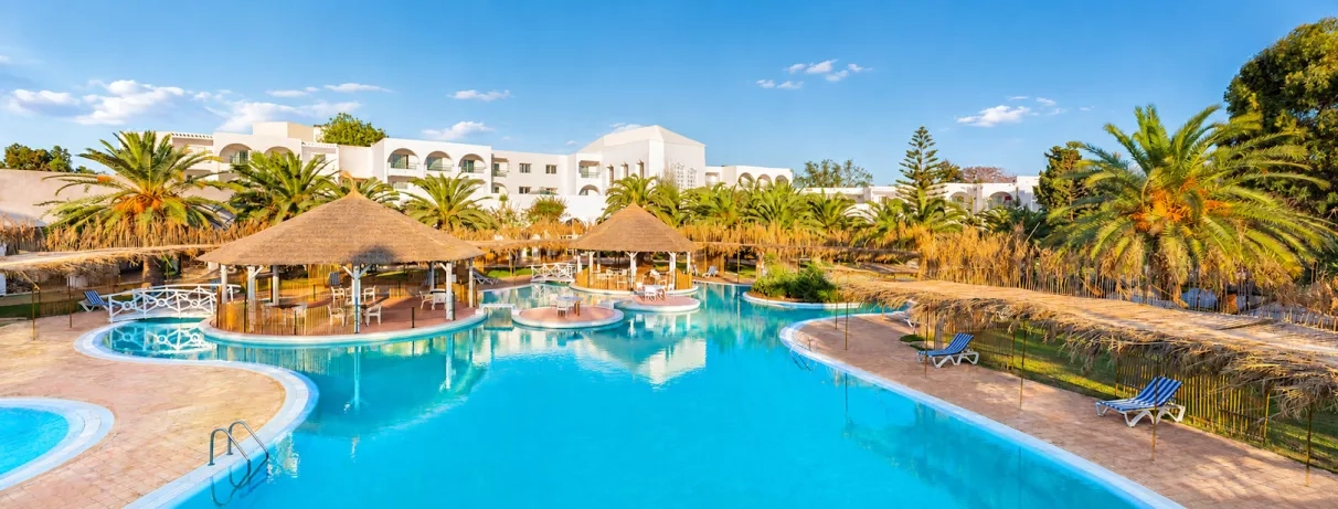 Grande piscine lagon du Mondi Club Shalimar Hammamet bordée de palmiers et d'aillottes en paille.