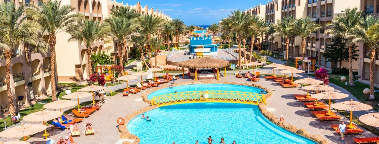 Grande piscine extérieure traversée par un pont en bois, entourée de bâtiments, parasols et palmiers à l'hôtel El Karma Aqua Beach.