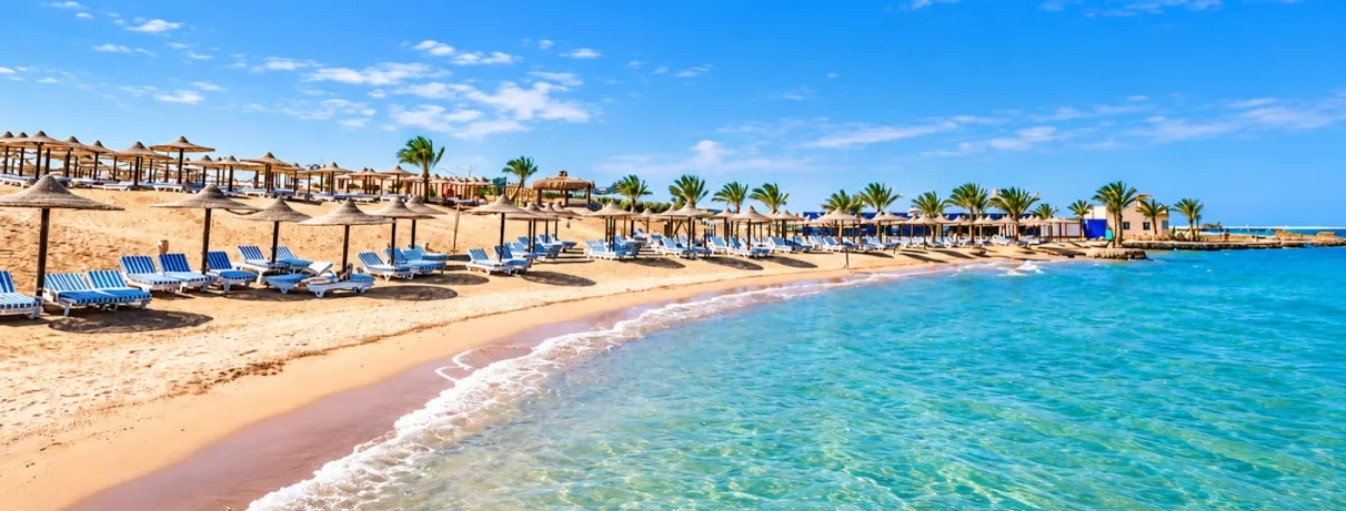 Plage privée de sable fin avec parasols en paille et transats au bord d'une eau turquoise à l'hôtel El Karma Aqua Beach Resort Hurghada.